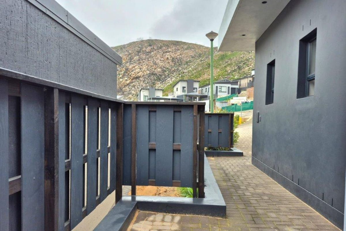 A paved side walkway featuring modern dark-toned timber privacy screens and a retaining wall, set against a backdrop of hillside residential properties.