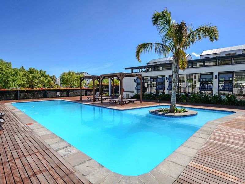 A well-maintained communal swimming pool area featuring timber decking, a central landscaped island with a palm tree, and a wooden pergola structure.
