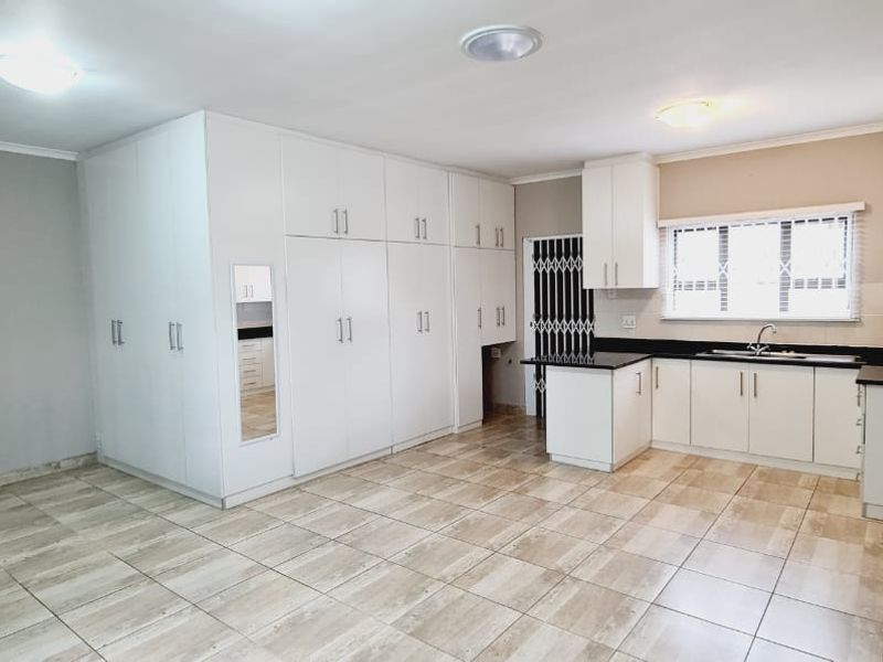 An open-plan living and kitchen area featuring extensive built-in white cabinetry, tiled flooring, and a functional kitchen layout with a sink and window.