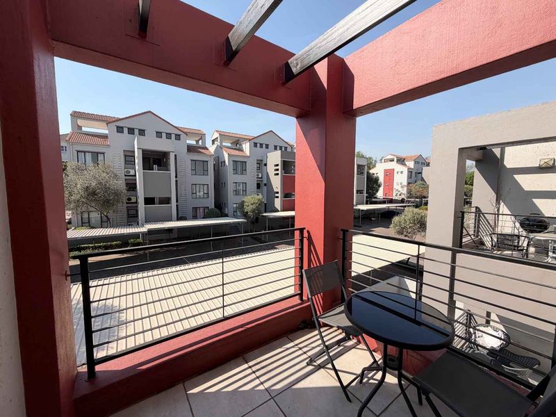 A covered balcony featuring a timber pergola, metal railings, and tiled flooring, overlooking the apartment complex grounds.