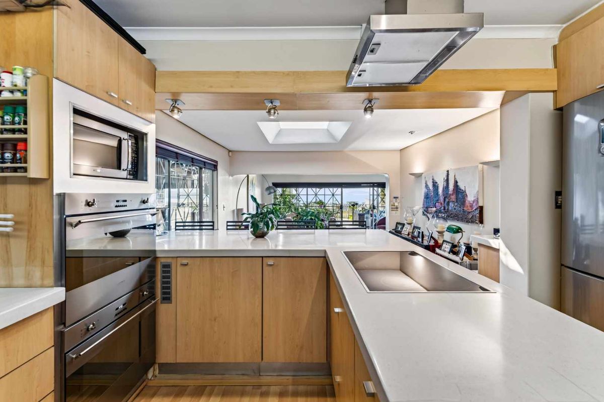 Modern kitchen featuring light wood cabinetry, stone countertops, built-in appliances, and a skylight, flowing into an open-plan living area with outdoor views.