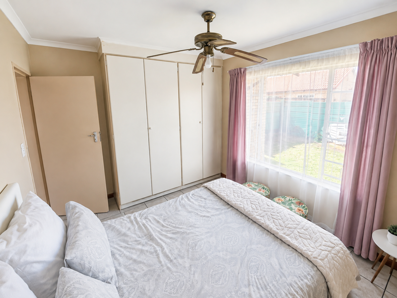 A well-lit bedroom featuring built-in cupboards, a ceiling fan, and large windows overlooking the rear garden area.