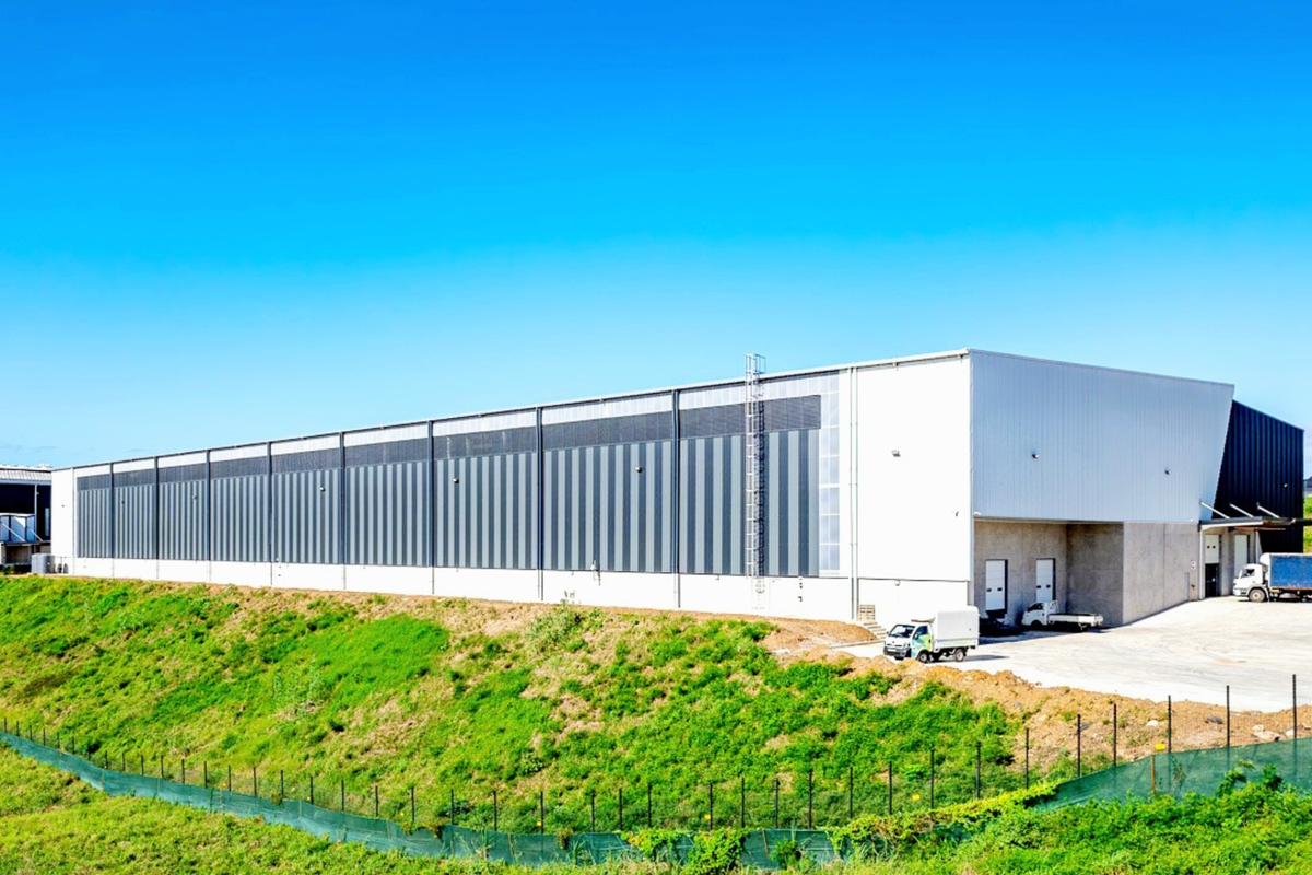 A large industrial warehouse facility featuring a modern metal facade, loading bays, and a concrete yard area, situated on a landscaped site.