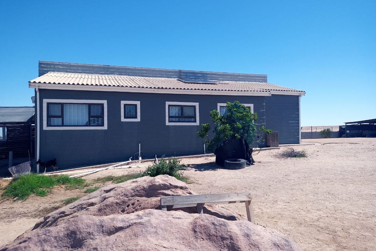 Rear view of a small holding dwelling featuring dark grey exterior walls, aluminium window frames with security bars, and roof-mounted solar panels.