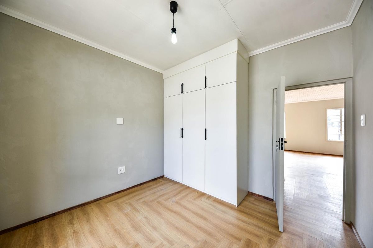 A bright, empty bedroom featuring light wood-look flooring, neutral walls, and a large, modern white built-in wardrobe with black hardware.