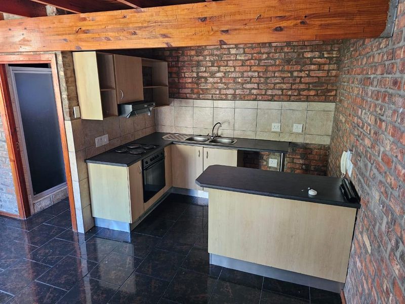 Compact kitchen featuring exposed brick walls, timber ceiling beams, dark tiled flooring, laminate cabinetry, and built-in cooking appliances.