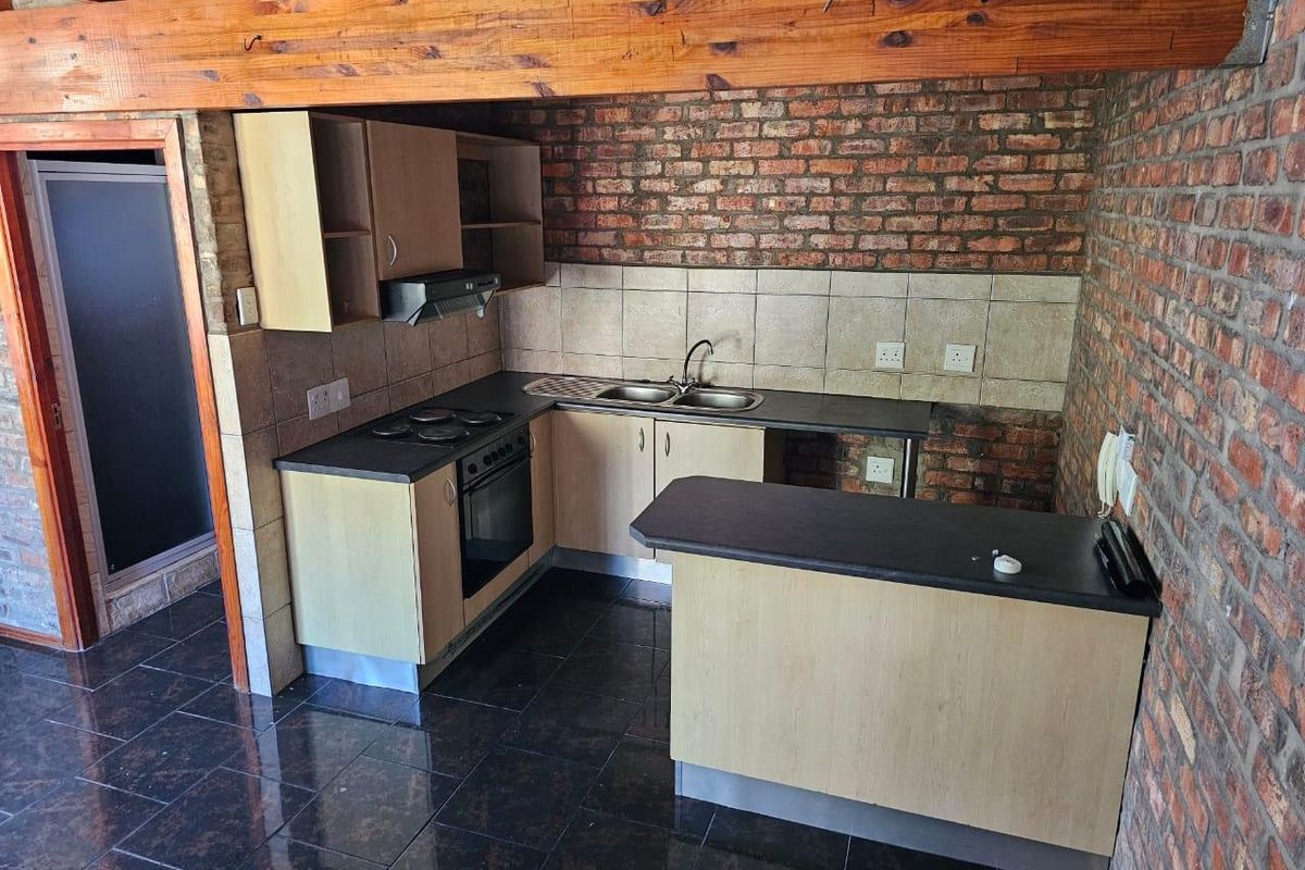 Compact kitchen featuring exposed brick walls, timber ceiling beams, dark tiled flooring, laminate cabinetry, and built-in cooking appliances.