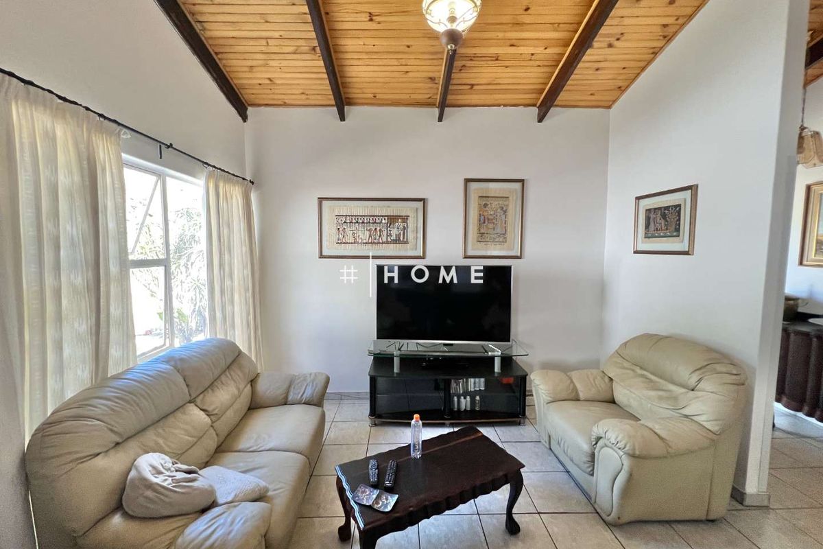 A living room featuring a vaulted timber-beamed ceiling, tiled flooring, and large windows allowing natural light.
