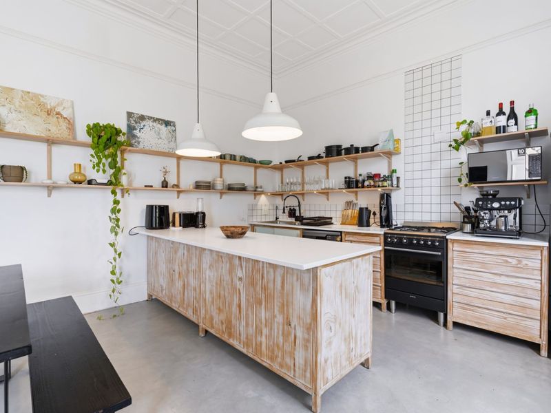 A bright kitchen featuring a central island, open timber shelving, a gas stove, and modern pendant lighting with a clean, minimalist aesthetic.