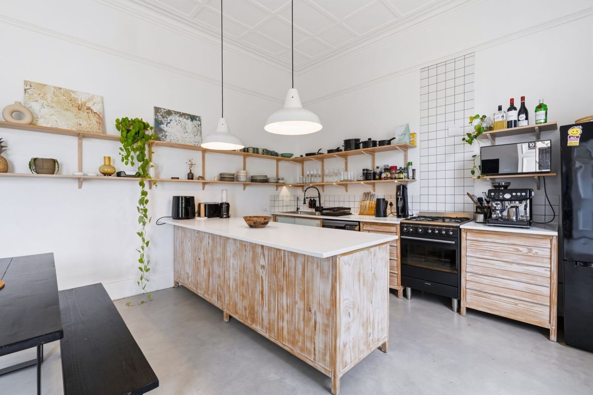 A bright kitchen featuring a central island, open timber shelving, a gas stove, and modern pendant lighting with a clean, minimalist aesthetic.