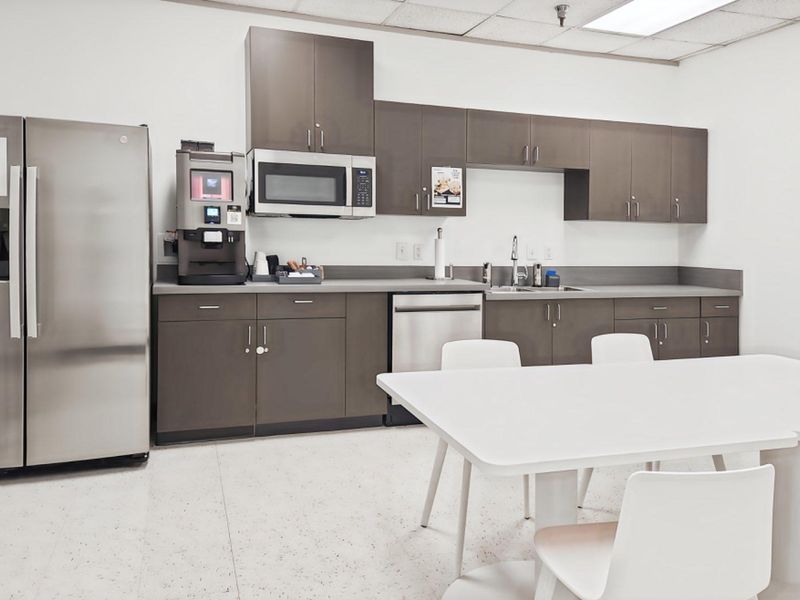 A functional office kitchenette featuring modern dark cabinetry, stainless steel appliances, a built-in microwave, and a drop ceiling with integrated lighting.