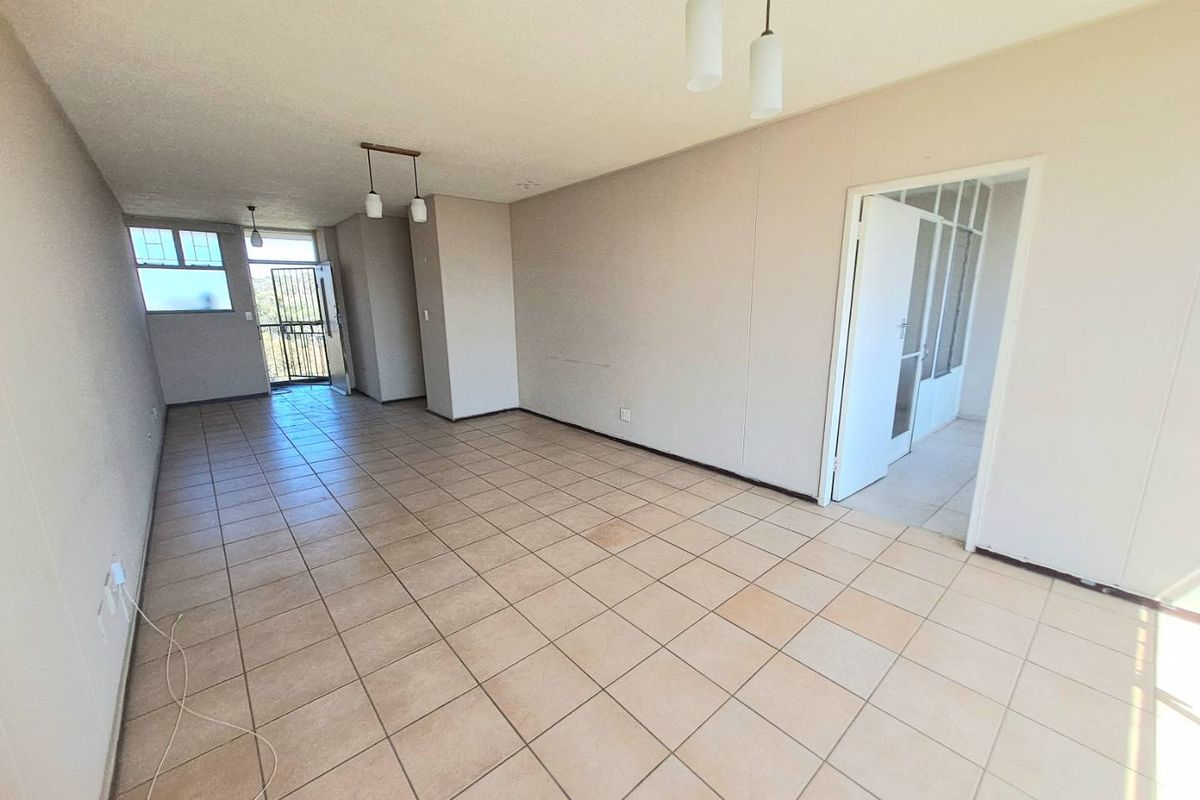 An empty, open-plan living area featuring neutral ceramic floor tiles, pendant lighting, and access to an exterior balcony through a security gate.