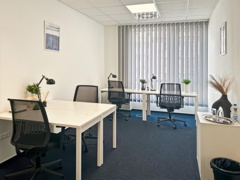 A professional office space featuring blue carpeting, a suspended ceiling with recessed lighting, and large windows fitted with vertical blinds.