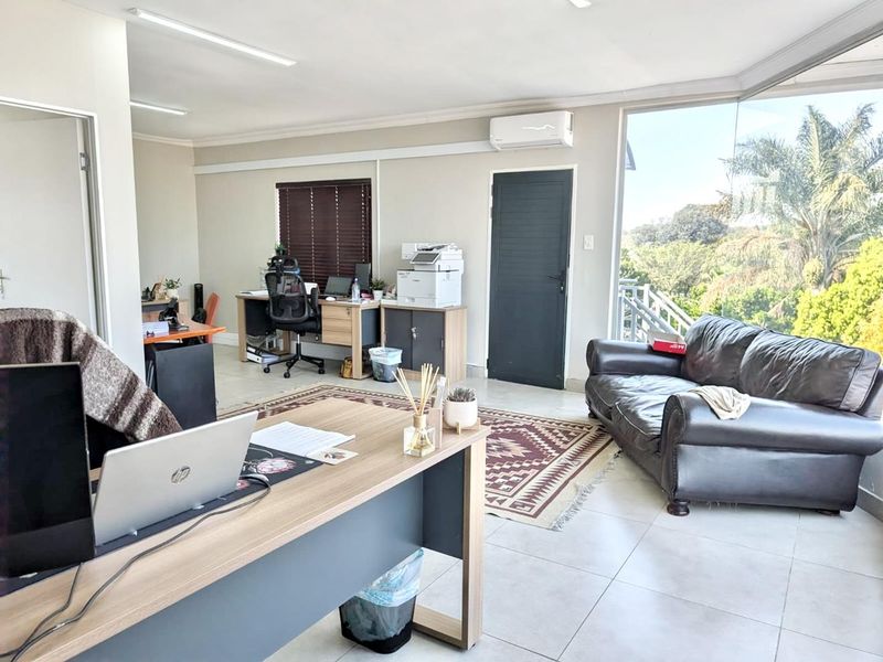 A bright and spacious office area featuring tiled flooring, an integrated air conditioning unit, and large windows providing natural light and garden views.