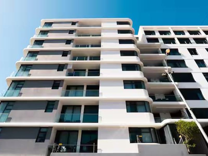 A low-angle view of a modern multi-story apartment building featuring clean white architectural lines, glass balcony balustrades, and large sliding glass doors.