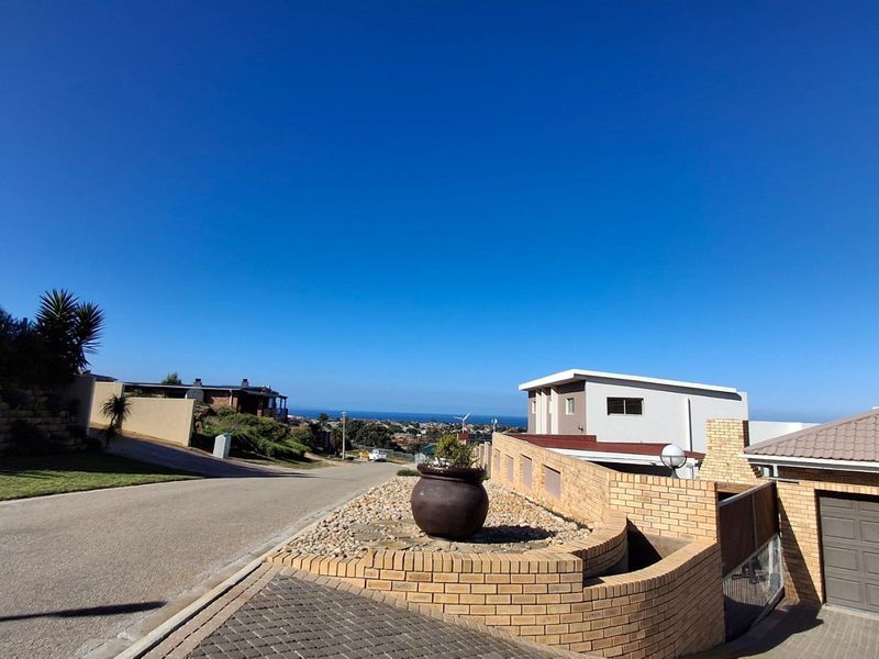 A street-level view of the property exterior, showcasing the paved driveway, brick boundary wall, and garage entrance with a distant coastal outlook.