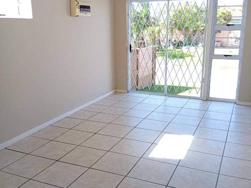 An empty living room featuring neutral tiled flooring, an air conditioning unit, and a sliding glass door with security gate leading to the garden.