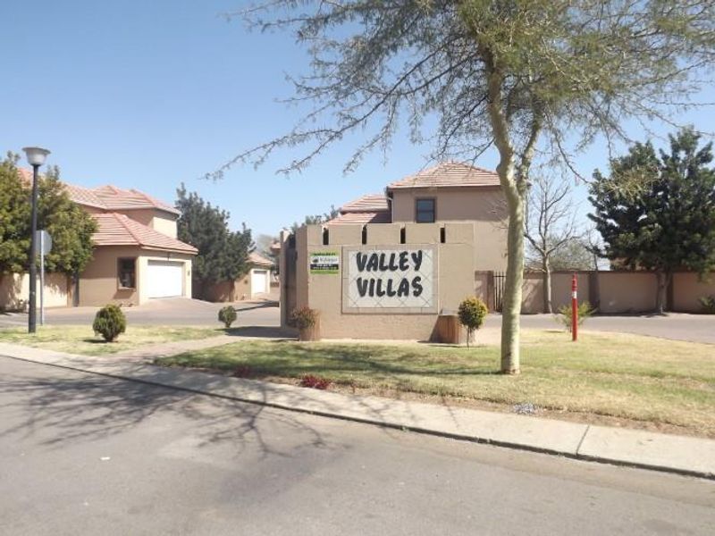 Street view of the Valley Villas townhouse complex entrance, featuring established landscaping, paved driveways, and residential units with tiled roofs.