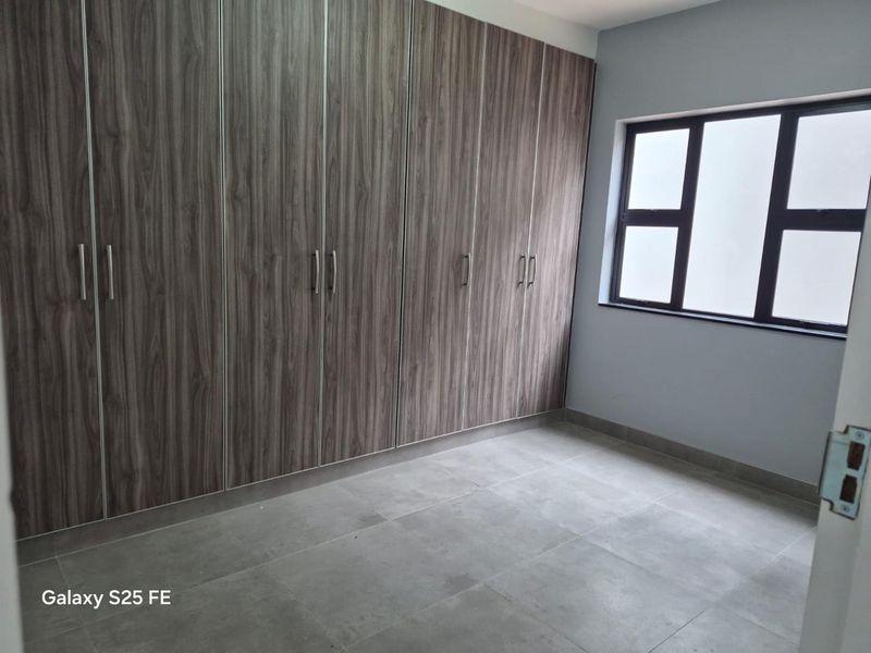 A clean, empty bedroom featuring modern wood-look built-in cupboards, neutral grey floor tiles, and a large aluminium-framed window.