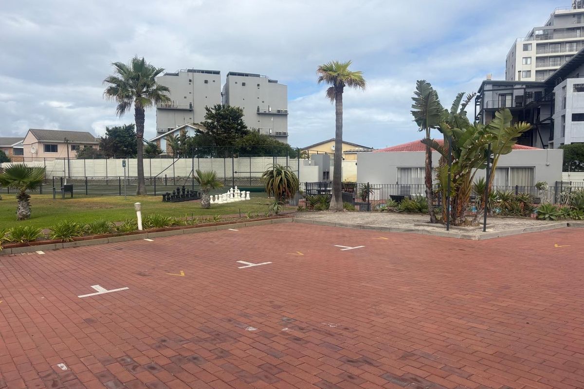 A paved parking area overlooking a communal garden space featuring a tennis court and landscaped grounds within the apartment complex.