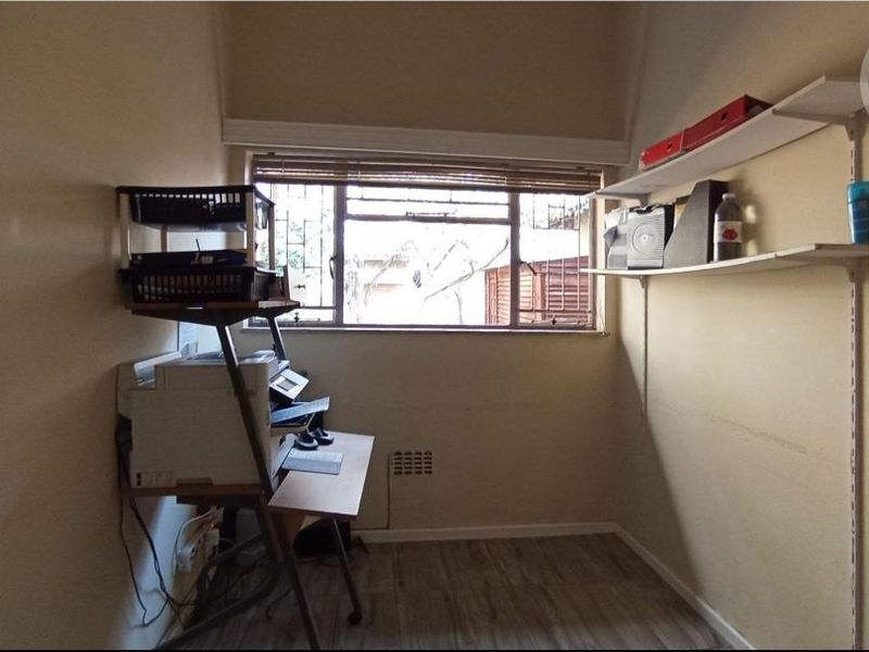 A compact home office space featuring laminate flooring, wall-mounted shelving, and a window with security bars.