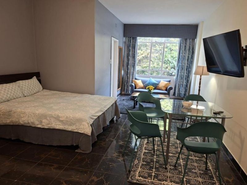 A spacious bedroom featuring dark tiled flooring, a large window with natural light, and a wall-mounted television.