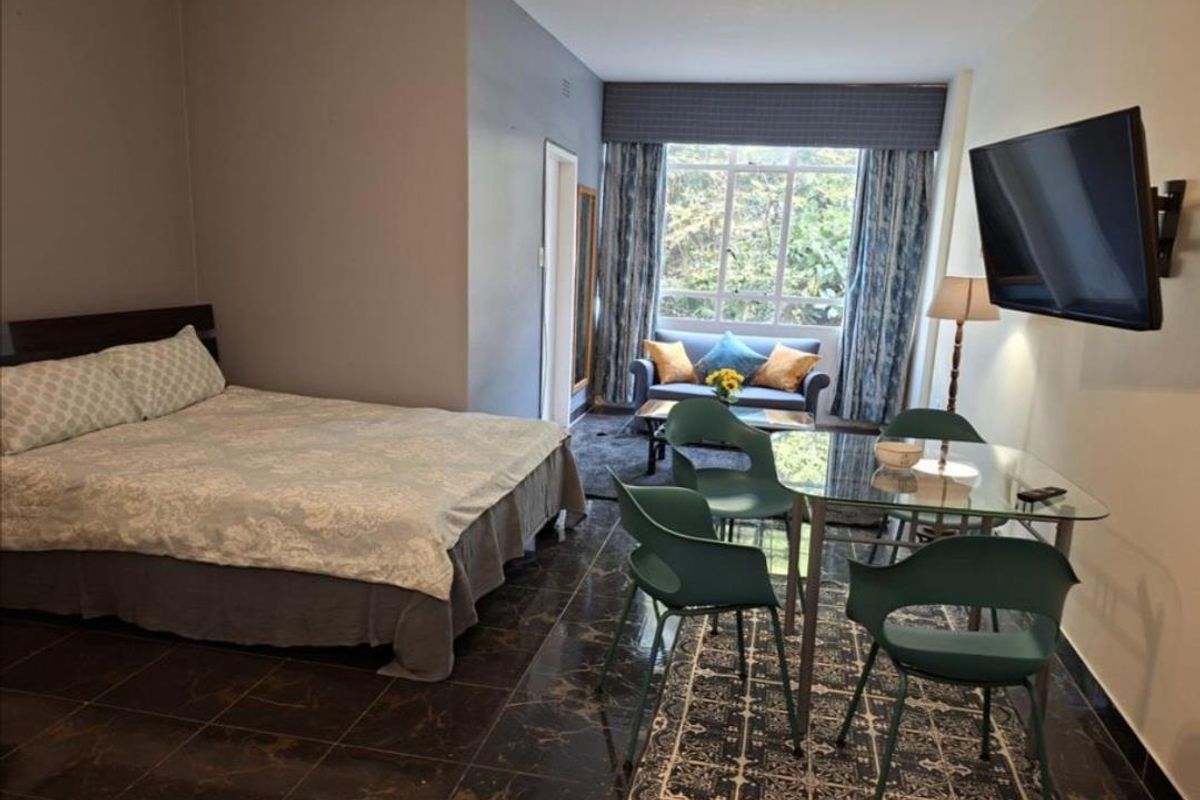 A spacious bedroom featuring dark tiled flooring, a large window with natural light, and a wall-mounted television.