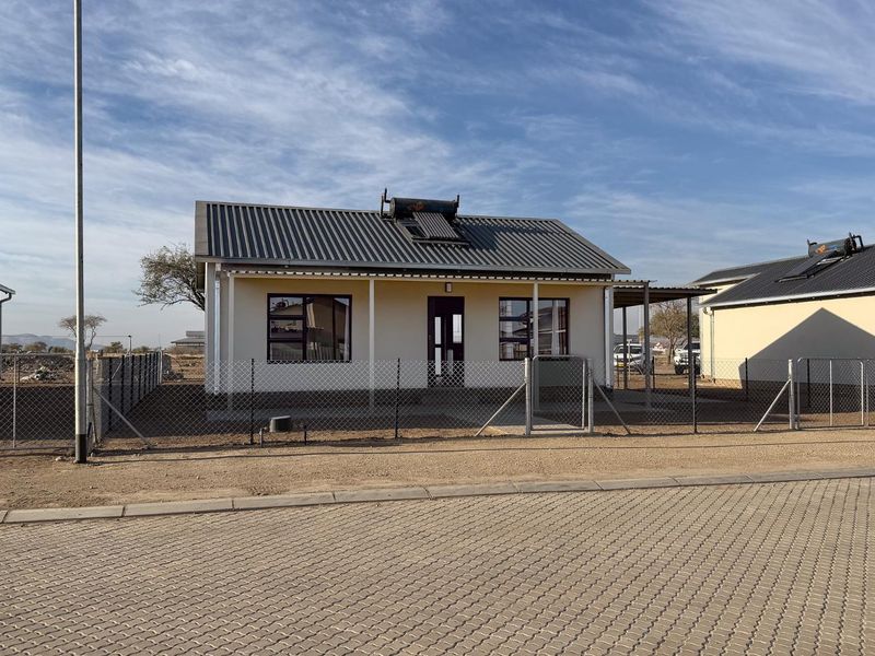 Front view of a single-story detached house featuring a corrugated metal roof, solar water heater, and a fenced yard with a paved driveway.