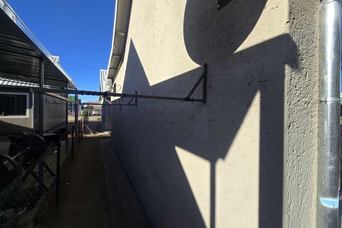 A side exterior view of the property featuring a paved walkway, wall-mounted clothesline, and metal downpipe along the building facade.