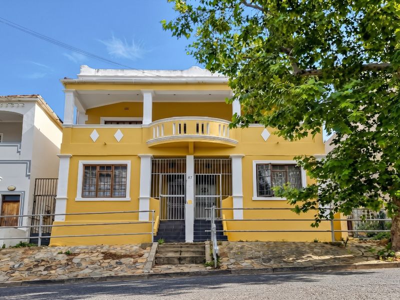 Front elevation of a yellow two-story apartment building featuring a balcony, security gates, and a stone retaining wall along the street frontage.