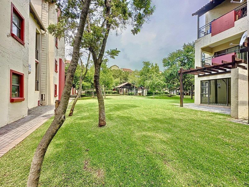 A well-maintained communal garden area featuring a lush lawn, mature trees, a timber pergola, and paved walkways between apartment blocks.