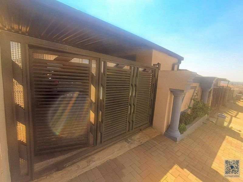 Front entrance view featuring a modern sliding metal gate, paved driveway, and decorative boundary wall pillars.