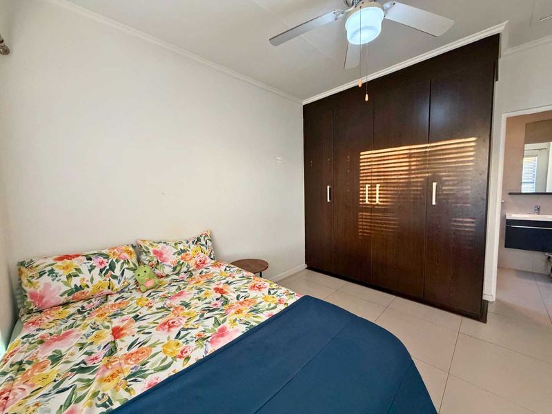 A well-maintained bedroom featuring dark wood built-in cupboards, a ceiling fan, and neutral tiled flooring, with an adjoining bathroom visible.