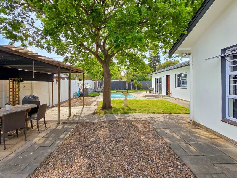 A spacious rear garden featuring a swimming pool, paved patio area, and a covered outdoor entertainment space under a mature tree.