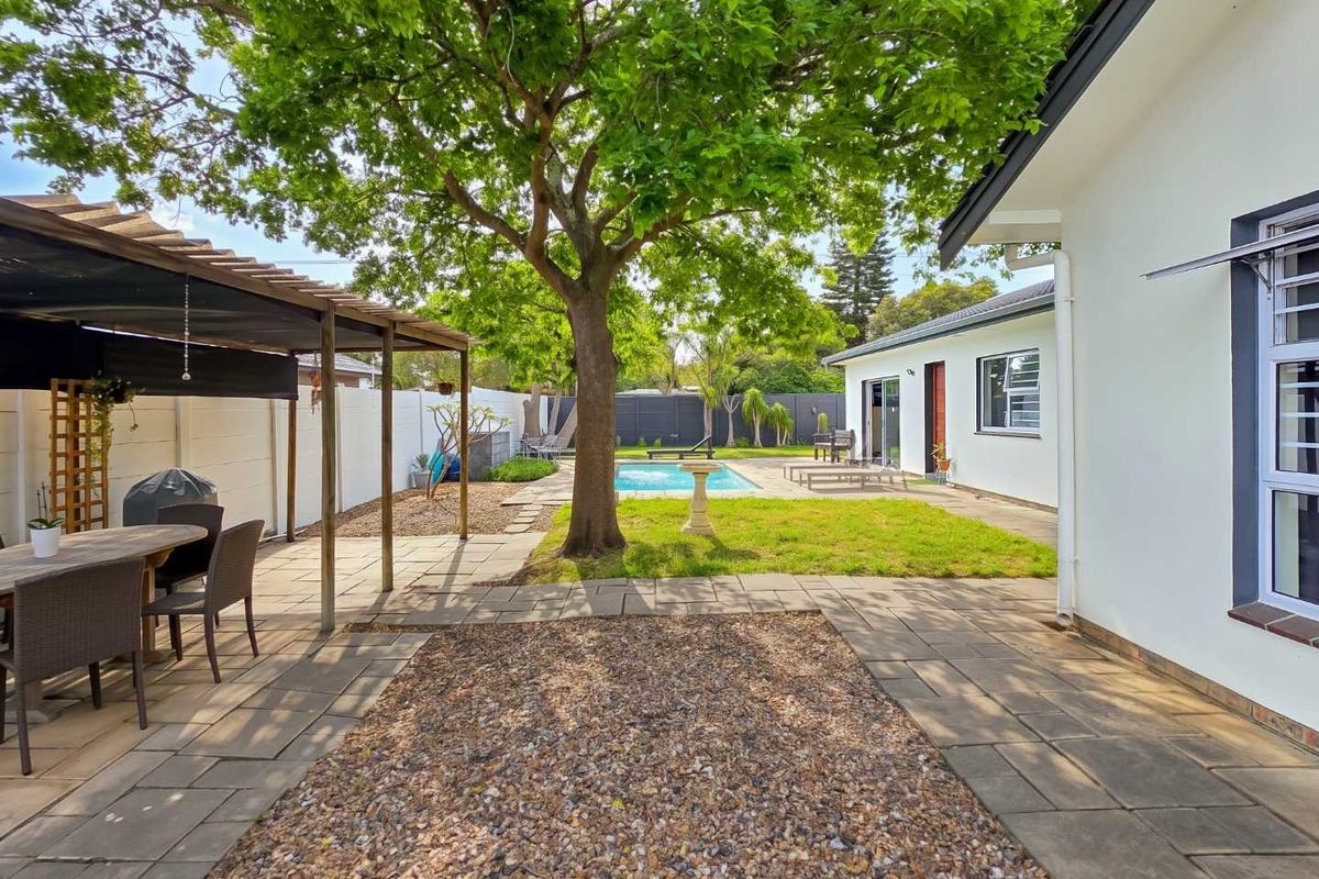 A spacious rear garden featuring a swimming pool, paved patio area, and a covered outdoor entertainment space under a mature tree.