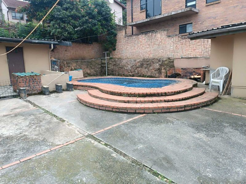 Outdoor pool area featuring a raised brick-edged swimming pool, concrete patio, and a built-in brick braai against a stone retaining wall.