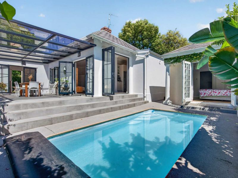 A well-maintained outdoor pool area featuring a paved deck, concrete steps, and a covered patio with a glass roof structure.