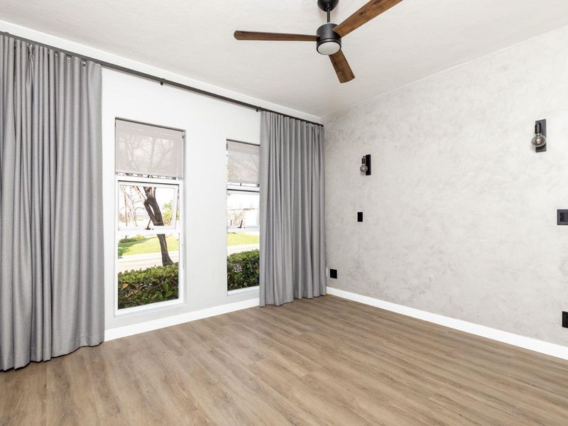 A bright, empty bedroom featuring modern laminate flooring, a ceiling fan, and a textured accent wall with contemporary wall sconces.