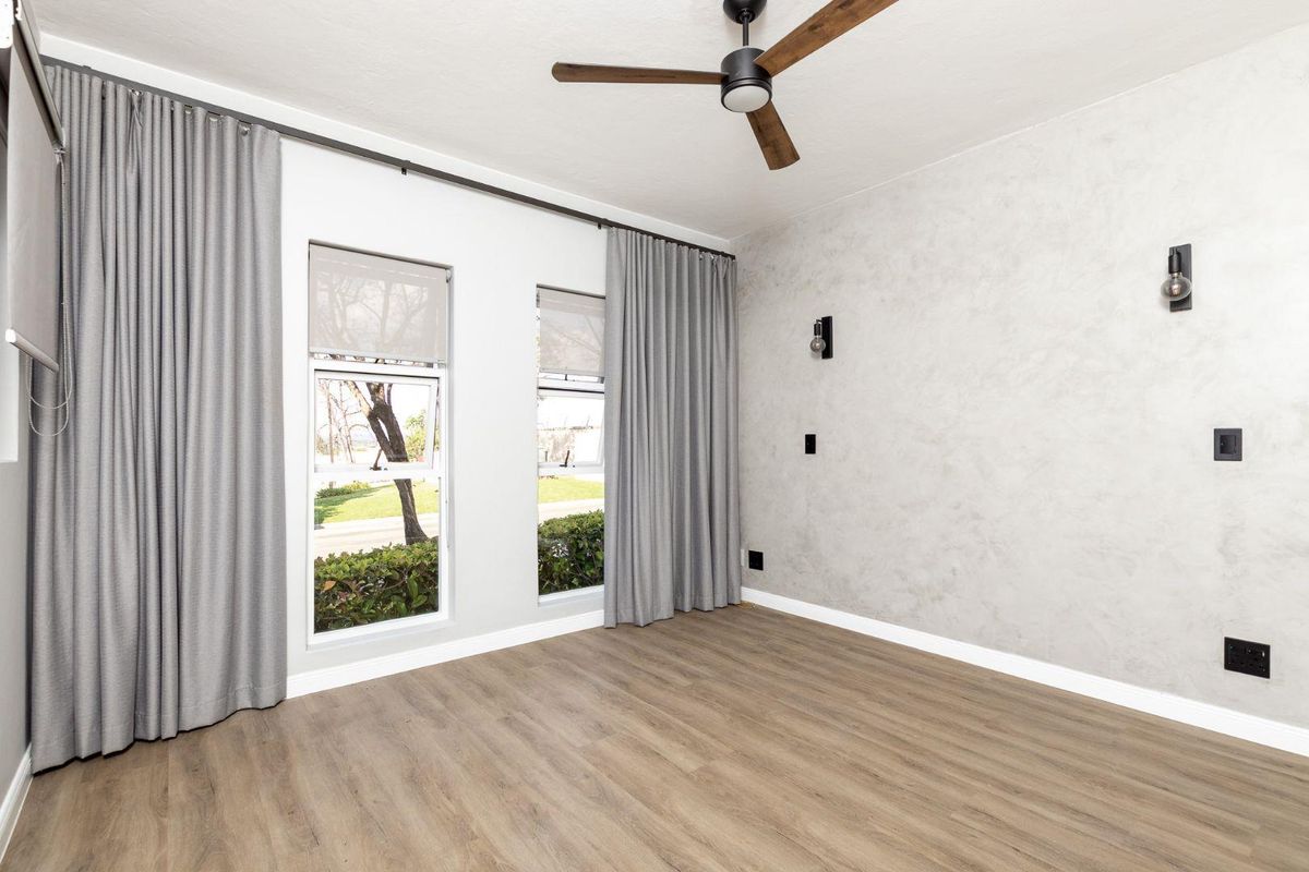 A bright, empty bedroom featuring modern laminate flooring, a ceiling fan, and a textured accent wall with contemporary wall sconces.