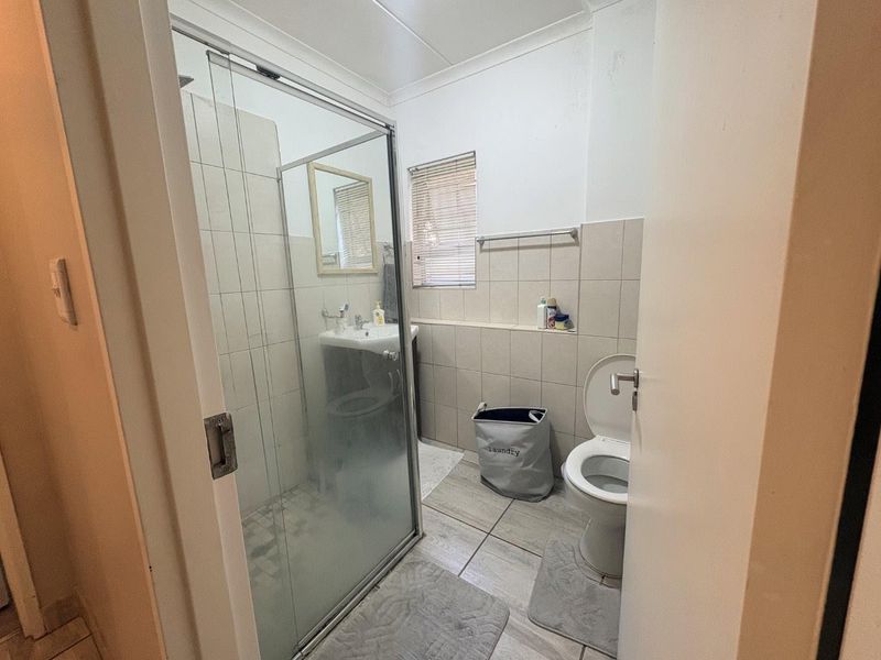 A functional bathroom featuring a glass-enclosed shower, pedestal basin, and toilet, finished with neutral wall and floor tiles.