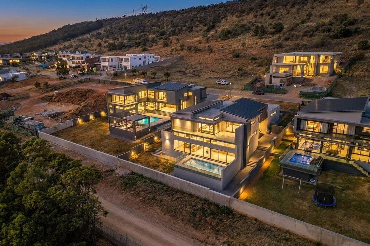 Aerial view of modern multi-level residential properties featuring private swimming pools, landscaped grounds, and secure boundary walls in a hillside estate setting.