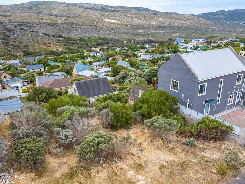 An elevated aerial view of a vacant residential plot featuring natural vegetation, overlooking a suburban neighborhood with mountain views in the background.