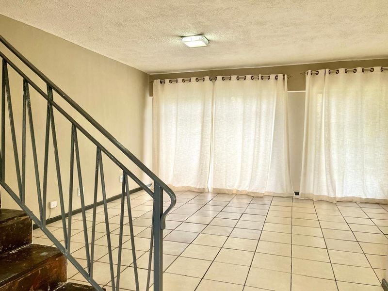 An empty living area featuring tiled flooring and a metal staircase railing, leading to large windows with curtain tracks.