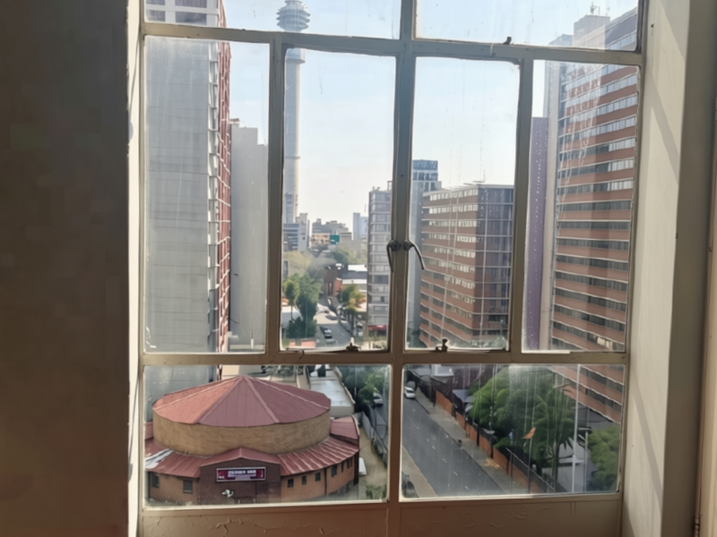 An urban city view captured through a large, multi-pane metal-framed window, showcasing surrounding high-rise buildings and street infrastructure.