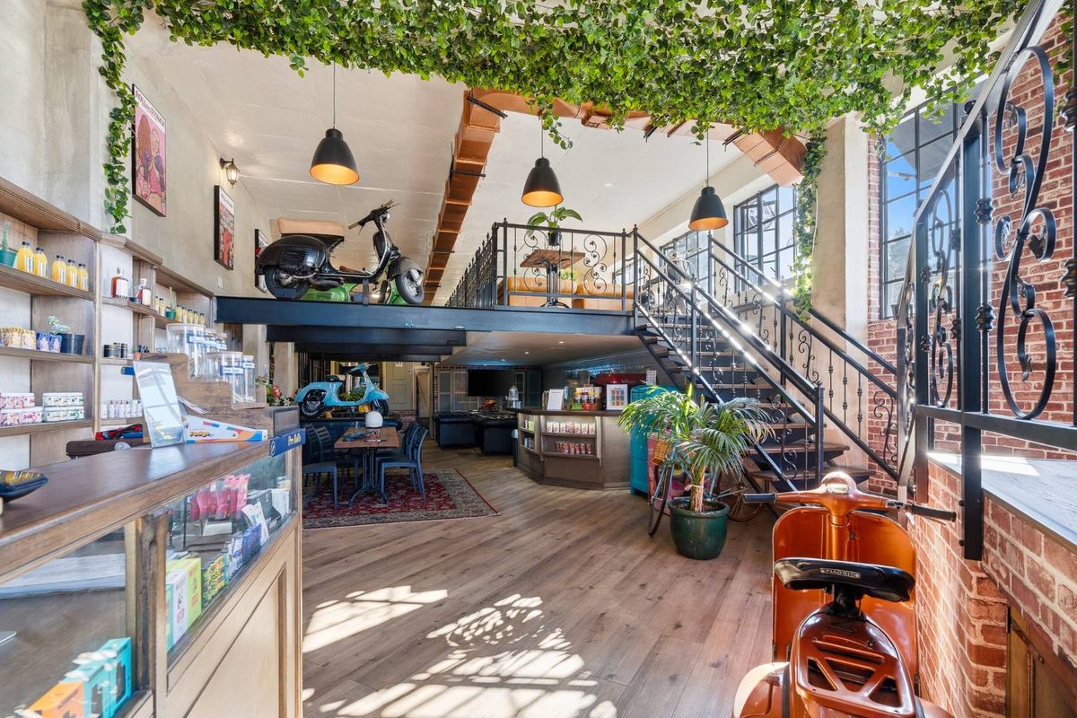 A spacious open-plan retail interior featuring a mezzanine level, exposed brickwork, industrial-style steel beams, and a prominent wrought iron staircase.