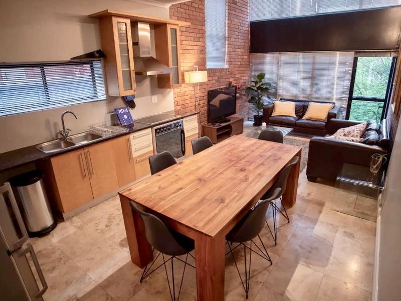 Open-plan living area featuring a modern kitchen with built-in cabinetry, stone flooring, and an exposed brick feature wall with large windows.