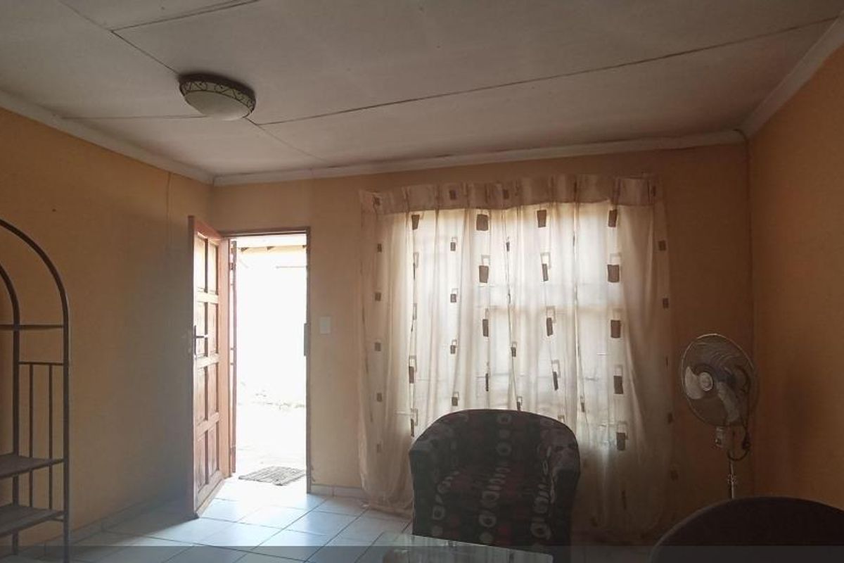 A living room area featuring tiled flooring, a wooden entrance door, and a large window with curtains, providing access to the exterior.