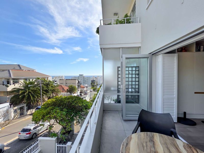 A bright, well-maintained balcony featuring tiled flooring, a metal railing, and a clear view of the surrounding neighborhood and distant ocean.