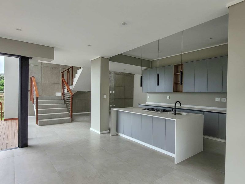 Modern open-plan kitchen featuring a central island with gas cooktop, sleek grey cabinetry, and seamless transition to an outdoor timber deck and staircase.