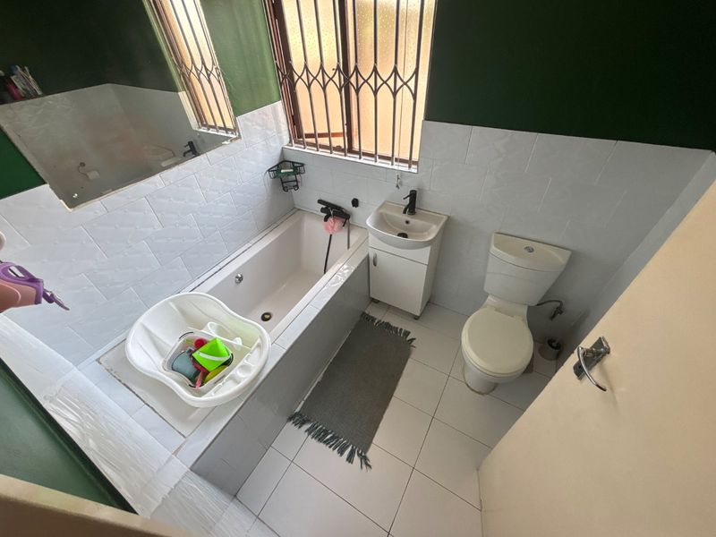 A functional bathroom featuring a built-in bathtub, vanity basin, and toilet, finished with white wall tiles and security bars on the window.